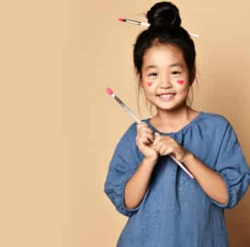 Cute smiling asian kid girl artist with painted red hearts on cheeks and with brush in hair and in hands over yellow wall background with copy space